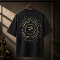 A black washed t-shirt hanging on a wooden hanger against a dark, atmospheric background with warm sunlight and shadows. The back of the shirt features a large distressed gold graphic with a central Om symbol and Sanskrit text.
