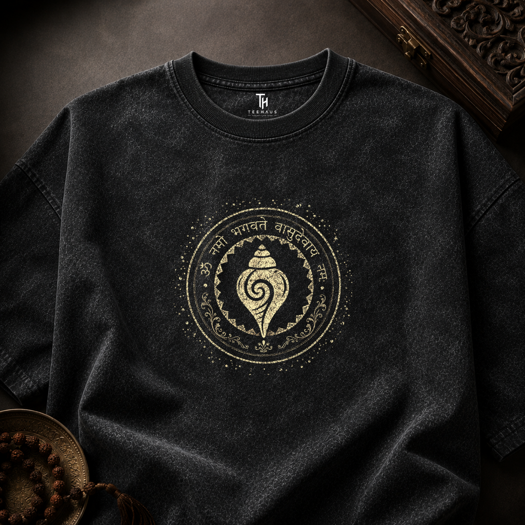 A dark charcoal gray crewneck t-shirt with a distressed wash, featuring a circular gold-toned graphic on the chest with a shell motif and Sanskrit text. The shirt is styled in a flat-lay arrangement on a dark textured background with decorative props.