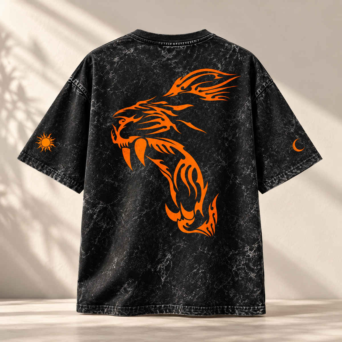 A black acid-wash t-shirt with a large orange tribal tiger graphic on the back, featuring smaller orange sun and moon graphics on the sleeves, displayed against a warm beige background with soft leaf shadows.