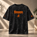 A black acid-wash graphic t-shirt with orange text in Devanagari script and claw marks on the front, displayed against a warm, sunlit textured wall.
