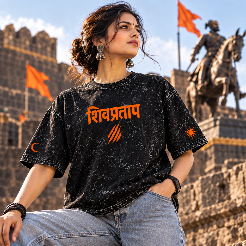 A woman wearing a black acid-wash graphic t-shirt with orange text and symbols, posing in front of a stone castle with orange flags and a statue.