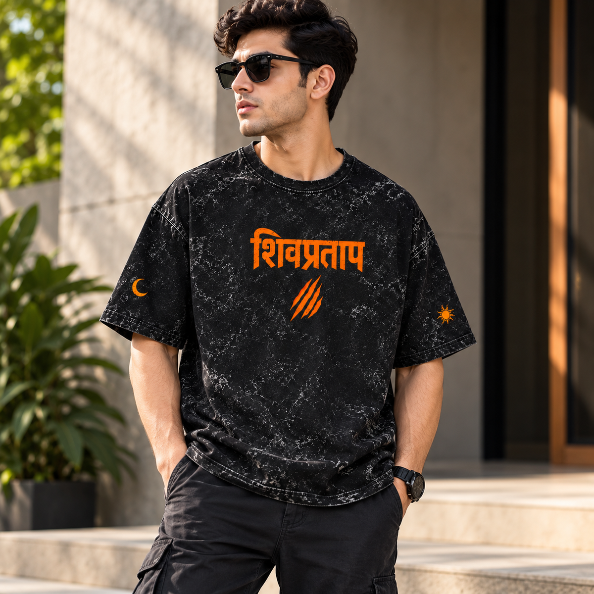 A man wearing a black short-sleeve t-shirt with a marbled pattern and orange text in Devanagari script on the chest, standing outdoors in front of a modern building.
