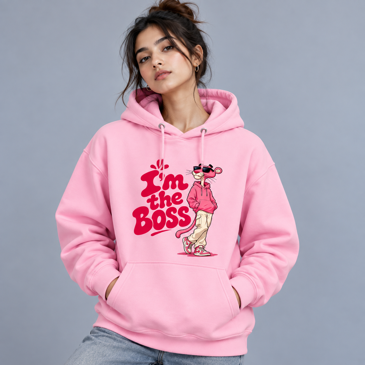 A woman wearing a light pink pullover hoodie with a graphic print of the Pink Panther character and the text 'I'm the Boss' in red and pink, paired with blue jeans against a solid gray studio background.