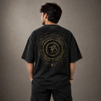 A man viewed from behind wearing a faded black t-shirt with a large gold circular spiritual graphic featuring an Om symbol and Sanskrit text on the back, set against a neutral gray studio background.