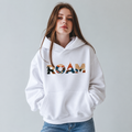 Woman wearing a white ROAM pullover hoodie with multicolor landscape lettering across the chest and a front pouch pocket.