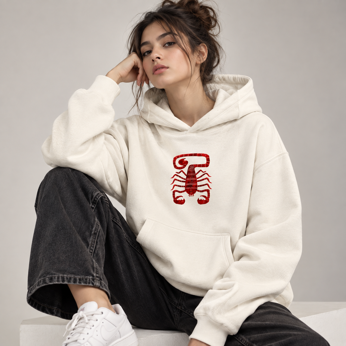 Seated woman wearing off-white Scorpio oversized hoodie with burgundy scorpion chest graphic, dark jeans and white sneakers.