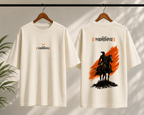 Two off-white short-sleeve T-shirts hanging on wooden hangers against a softly lit wall with leaf shadows. The front shirt has a small orange and black chest graphic with Devanagari text, and the back shirt has a large black rider graphic over a bold orange brushstroke with matching Devanagari text above.