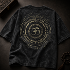 A dark gray mineral-wash t-shirt with a large gold circular graphic featuring an Om symbol and Sanskrit text, displayed flat on a dark textured surface with decorative props.