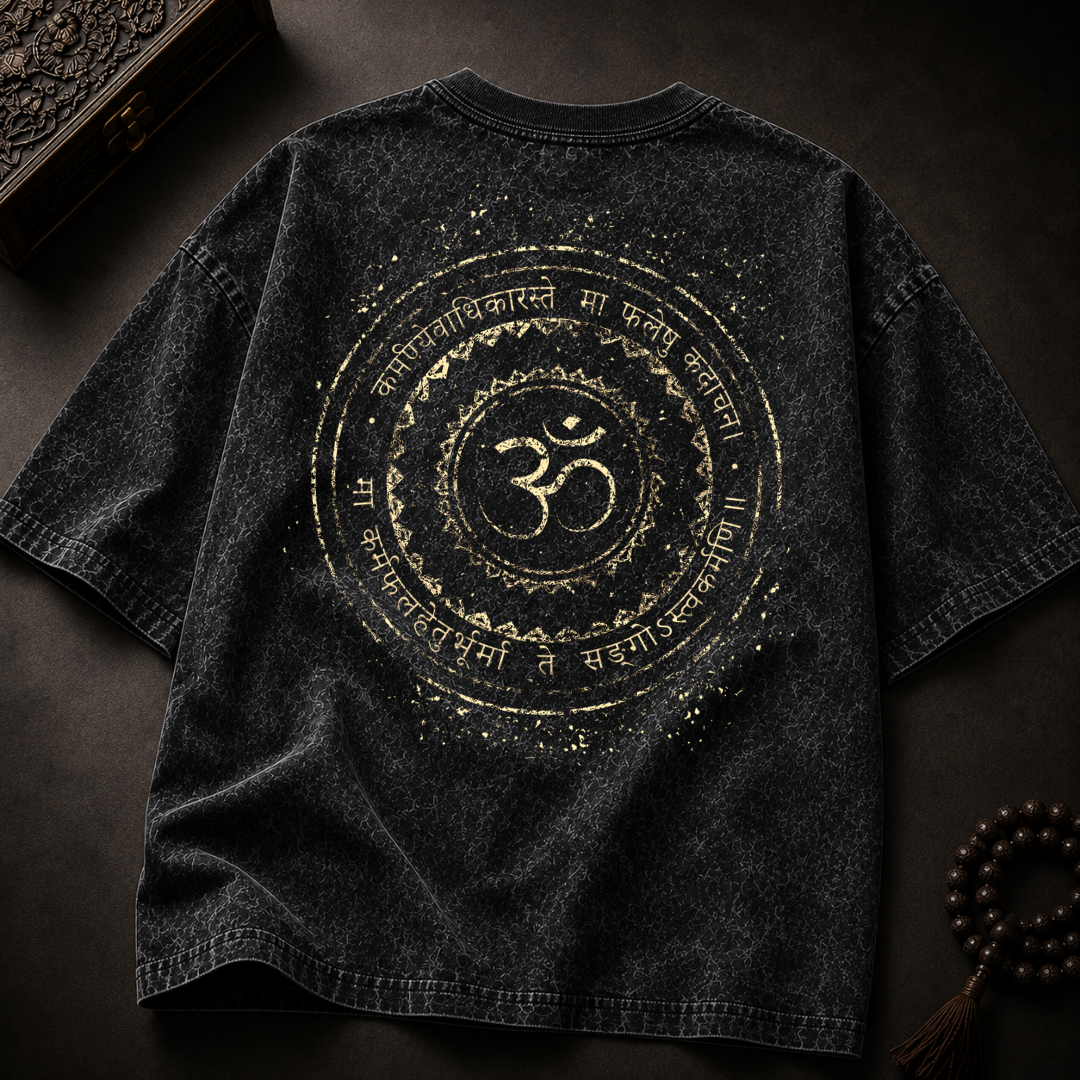 A dark gray mineral-wash t-shirt with a large gold circular graphic featuring an Om symbol and Sanskrit text, displayed flat on a dark textured surface with decorative props.